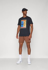 Μαύρο μπλουζάκι με πολύχρωμο γραφικό σχέδιο; καφέ σορτς; λευκά αθλητικά παπούτσια; και μαύρο καπέλο. Το μοντέλο στέκεται μπροστά σε ουδέτερο φόντο.