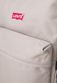 Béžový látkový batoh s přední kapsou, na níž se nachází červené logo Levi's. Viditelný je texturovaný materiál a zpevněné švy.