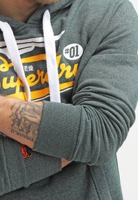 Hombre vistiendo una sudadera gris de Superdry con cordones blancos, con los brazos cruzados que muestran un antebrazo tatuado y detalles del logo visibles.