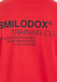Rød bomulds t-shirt med stor sort tekst, der står "SMILODOX TRAINING CLUB" og motiverende sætninger som "STOL PÅ PROCESSEN" og "BLIV FOKUSERET."