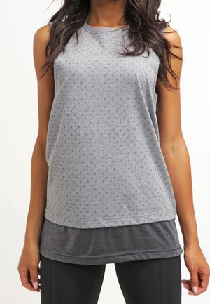 Kobieta ubrana w szary, bezrękawnikowy, warstwowy top do ćwiczeń oraz czarne legginsy na jednolitym białym tle.