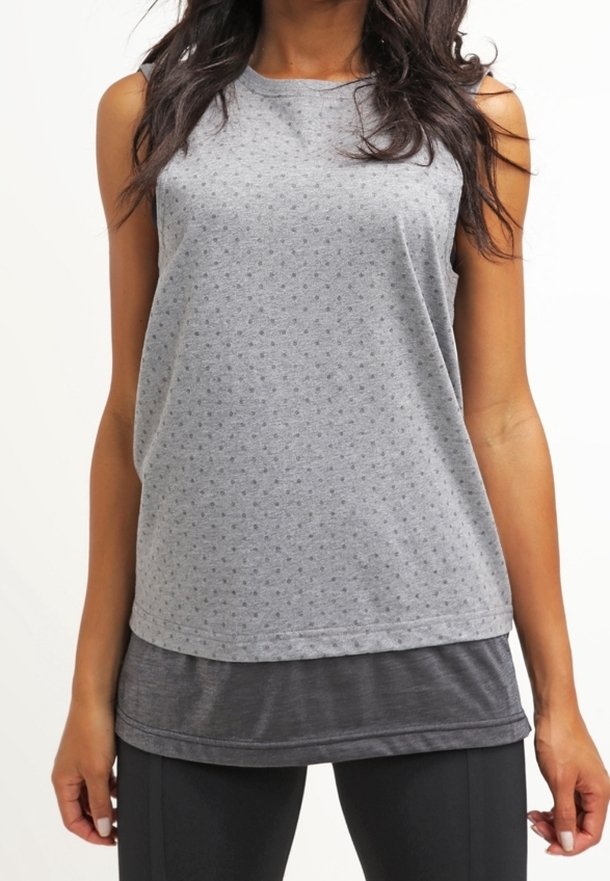 Mujer con camiseta deportiva sin mangas gris de capas y leggings negros frente a un fondo blanco liso.