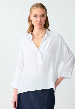 Femme portant un chemisier blanc à manches longues avec un col en V et un collier de perles bleu, associé à une jupe en jean foncé, posant avec la main sur la hanche.
