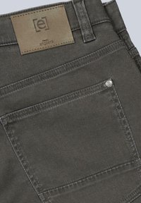 Graue Denim-Hose mit einem braunen Lederetikett mit eingeprägtem Logo, dezenten Stichelementen und einem kleinen Metallknopf auf der hinteren Tasche.