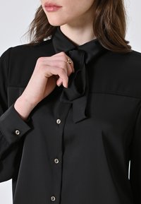 Schwarze Bluse mit gebundenem Kragen, glatter Satin-Oberfläche und silbernen Knopfdetails an der Vorderseite. Die Ärmel sind an den Manschetten geknöpft.