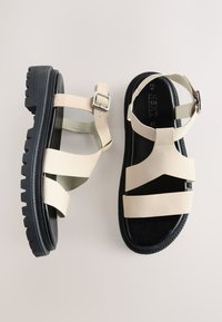 Sandale din piele bej cu multiple curele late, cataramă reglabilă la gleznă și talpă groasă din cauciuc negru cu un model robust de crampoane.