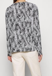 Sort-hvid bluse med lange ærmer, der har et tekstureret, abstrakt mønster, en afslappet pasform og rund halsudskæring. Kombineret med lyse bukser.