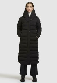 Cappotto lungo imbottito nero con colletto alto e chiusura a bottoni, caratterizzato da cuciture diagonali e due tasche laterali. Indossato sopra pantaloni scuri e stivali.