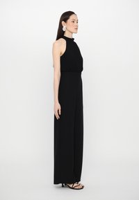 Svart, ermeløs jumpsuit med høy hals og avslappet passform, laget av mykt stoff, med vide ben og minimalistisk design.