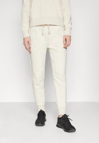 Pulover din tricot bej cu manșete și tiv fin din jerseu, asortat cu pantaloni de trening tapered, având talie elastică și șnur, purtat cu adidași negri.