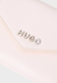 Lys rosa teksturert lommebok med et sølvfarget "HUGO"-logo. Lommeboken har et trekantet design med sømmer og en jevn overflate.
