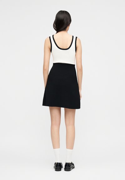 Γυναίκα με μακριά σκουρόχρωμα μαλλιά, φορόντας λευκό αμάνικο τοπ με μαύρη επένδυση, μαύρη φούστα, λευκές κάλτσες και μαύρα παπούτσια, στέκεται γυρισμένη προς τα πίσω.