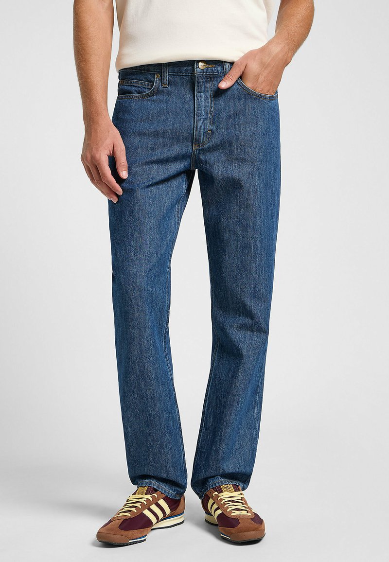 Pantalones vaqueros de mezclilla azul con un corte recto, cinco bolsillos y cierre de botón dorado. Llevados con zapatillas deportivas marrones y amarillas.