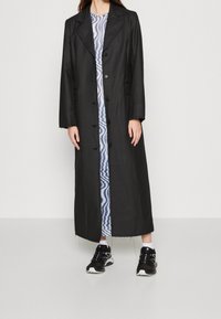 Manteau long noir avec un col structuré, des boutons à l'avant et des détails de coutures visibles, porté sur une robe bleue à motif et des baskets noires.