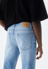 Jeans in denim azzurro chiaro con finitura testurizzata, caratterizzati da una toppa in pelle marrone con la scritta "PULL&BEAR" sulla cintura posteriore e cinque tasche.