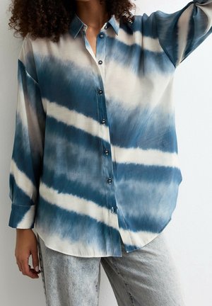 Mujer vistiendo una camisa abotonada de manga larga con estampado tie-dye azul y blanco y jeans de lavado claro, frente a una pared lisa.