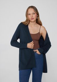 Cárdigan azul marino de material suave con mangas largas y un diseño abierto. Llevado sobre una camiseta de tirantes marrón y combinado con unos jeans azules.