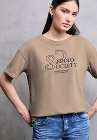 Béžové tričko vyrobené z měkké bavlny, se černým grafickým potiskem "STUDIO. SOCIETY." v uvolněném střihu a s ohrnutými krátkými rukávy. V kombinaci s modrými džíny.