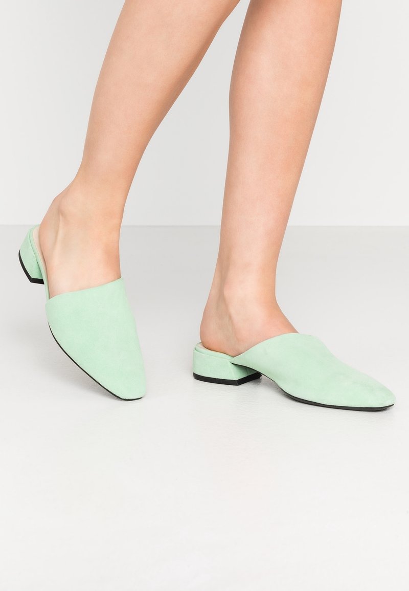 Jambes portant des mules vert pastel à talon bas et bouts pointus sur un fond neutre clair.