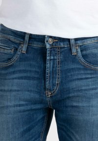 Dunkelblaue Jeans aus Denim mit einer leicht verblassten Textur, ausgestattet mit einem Reißverschluss, vorderen Taschen und kontrastierenden gelben Steppnähten.