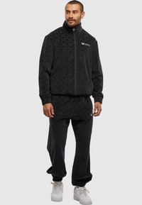 Homme portant une veste zippée texturée noire et un pantalon assorti, avec des baskets blanches, se tenant devant un fond neutre.