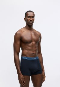 Black cotton boxer briefs with a blue Calvin Klein waistband. Smooth texture, elastic waistband, fitted design. Model stands with arms relaxed.