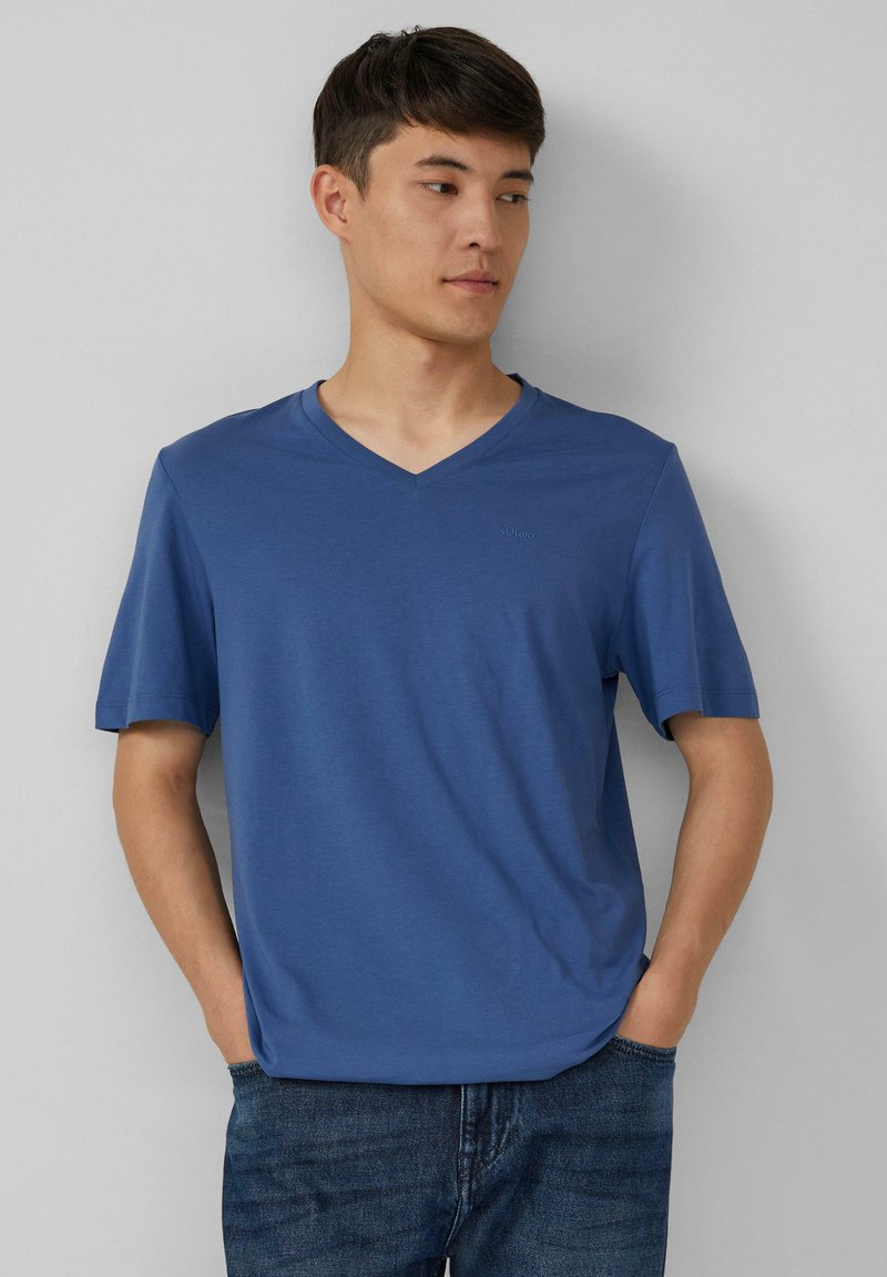 Niebieska koszulka z dekoltem w serek, wykonana z miękkiego materiału, z krótkimi rękawami i subtelnym logo na piersi. Zestawiona z dżinsami.