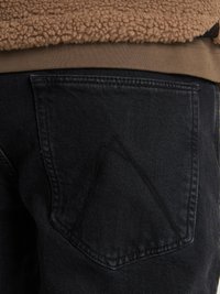 Jeans de mezclilla negra con un diseño distintivo de bolsillo trasero triangular. La tela tiene una textura suave, complementada por una parte superior de forro polar marrón.