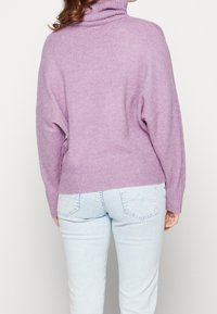 Frau, die einen lavendelfarbenen Rollkragenpullover und hellblaue Jeans trägt, von hinten vor einem schlichten weißen Hintergrund gezeigt.