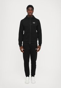 Črna zip-up kapuca z nastavljivo vrvico, usklajene trenirke in bele logotipne akcenti. Narejeno iz teksturirane tkanine. Enostavni beli športni copati.