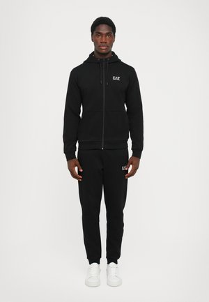 Haut à capuche noir zippé avec cordon de serrage réglable, pantalon de jogging assorti et accents de logo blancs. Fabriqué en tissu texturé. Baskets blanches unies.