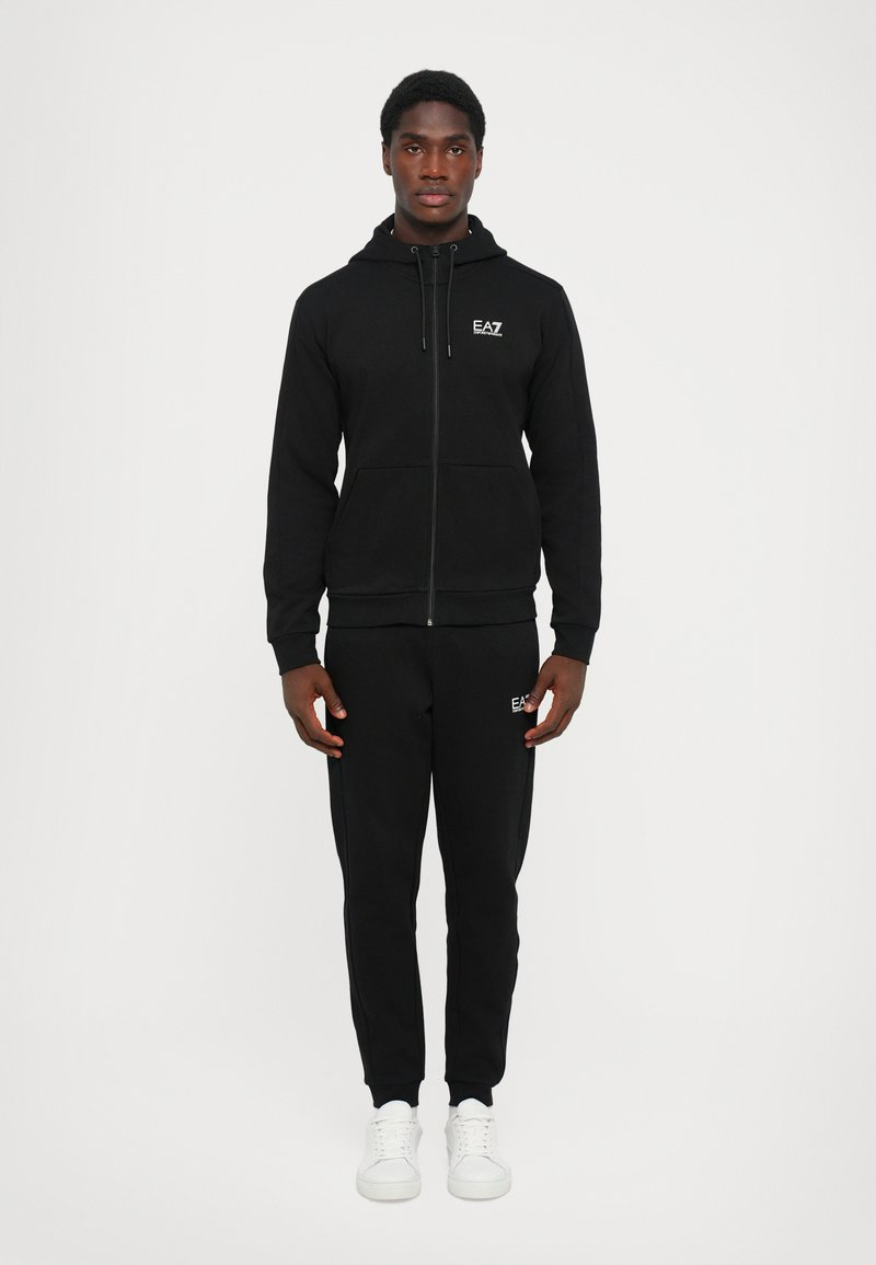 Svart hettegenser med glidelås og justerbar snor, matchende sweatpants, og hvite logo-detaljer. Laget av strukturert stoff. Enkle hvite joggesko.