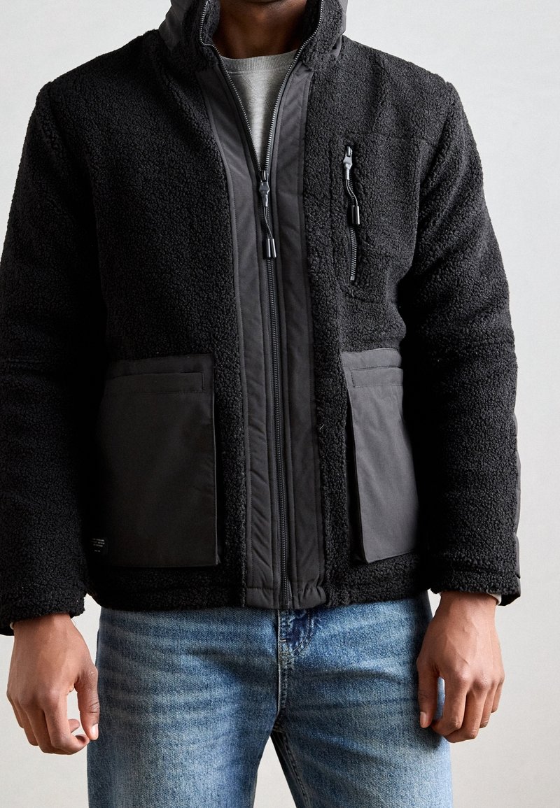 Personne portant une veste polaire noire avec des poches zippées sur le devant et la poitrine, associée à un jean bleu, sur un fond uni.