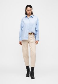 Mujer con camisa azul claro oversize, pantalones beige con cinturón marrón y botas negras hasta el tobillo, de pie frente a un fondo blanco liso.