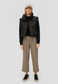 Schwarze Steppweste mit hohem Kragen, strukturierter schwarzer Strickpullover, karierten beige Hosen und schwarzen Plateausneakern.