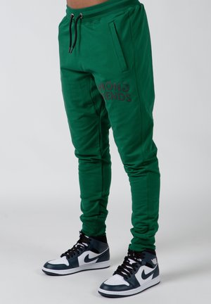 Green joggers made of soft fabric, featuring a slim fit, side pockets, and "WRONG FRIENDS" printed in dark text on the left thigh.