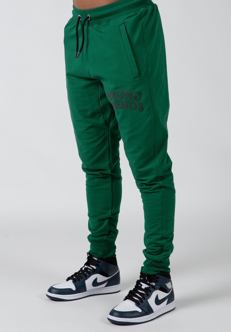 Groene joggers van zacht materiaal, met een slim fit, zijzakken en "WRONG FRIENDS" in donkere tekst op de linker bovenbeen gedrukt.