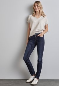 Beige stickad kortärmad v-ringad topp i kombination med mörkblå skinny jeans och vita sneakers. Outfiten har en avslappnad passform med en subtil struktur.