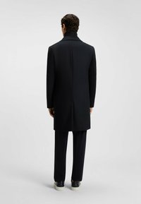 Schwarzer Wollmantel mit hohem Kragen, geradem Schnitt und Rückenschlitz; getragen über dunklen Hosen und schwarzen Sneakers. Glatte Textur.