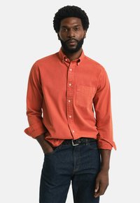 Rostorange Hemd mit Knopfleiste, einer Brusttasche und hochgekrempelten Ärmeln, kombiniert mit dunkelblauen Jeans und einem geflochtenen schwarzen Gürtel.
