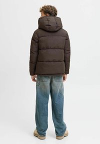 Brūns pūkains zābaks ar kapuci, kas piedāvā horizontālu quilted dizainu, un apvienots ar izbalējušām ziliem platām biksēm un bēšiem apaviem.