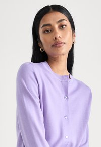 Lavendelkofta med rund hals, knäppning och långa ärmar. Tillverkad av mjukt tyg med en slät yta och subtil lyster.