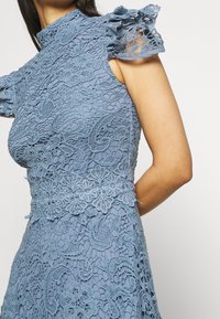 Niebieska sukienka koronkowa z wysokim dekoltem, krótkimi falbaniaste rękawami i skomplikowanymi wzorami kwiatowymi. Posiada dopasowaną talię z koronkowymi detalami.