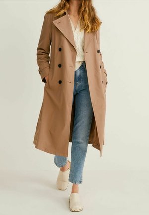 Vrouw lopend met een beige dubbelgeknoopte trenchcoat, witte gebreide trui, blauwe spijkerbroek en witte klompen tegen een effen achtergrond.