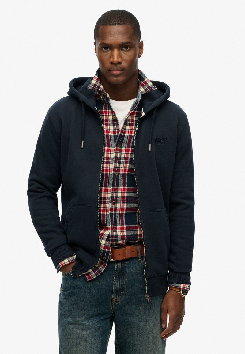 Marineblå zip-up hoodie lavet af blødt stof. Bæres over en rød og blåternet skjorte med knapper. Har en kengurulomme og hætte med snøre.
