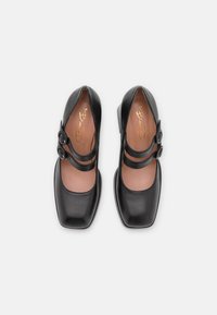 Black leather shoes with a square toe, two round buckle straps, and a low heel. The insole features a gold logo. Smooth texture.