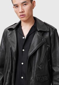 Schwarze Leder-Bikerjacke mit aufgeschlagenem Revers, mehreren Reißverschlusstaschen und Nietenakzenten. Trägt man über einem schwarzen Hemd mit Kragen.
