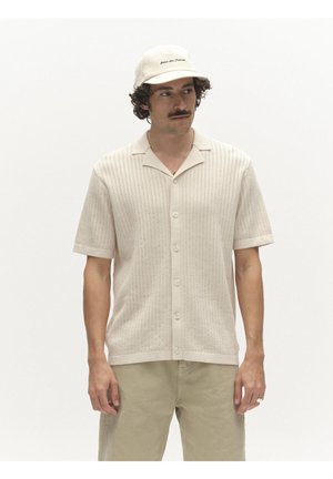 Férfi bézs kötött rövid ujjú, gombos felsővel, hozzáillő bézs nadrággal és krémszínű sapkával, amelyen felirat található az elején, fehér háttér előtt áll.