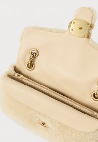 Bēša rokassoma ar krēmkrāsas kažokādu, zelta metāla detaļām, rāvējslēdzēja kabatu un ķēdes akcentiem. Mīksta tekstūra un strukturēts dizains.