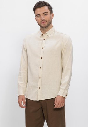 Man staand met een beige lange mouwen blouse met knopen en een bruine broek tegen een effen witte achtergrond.